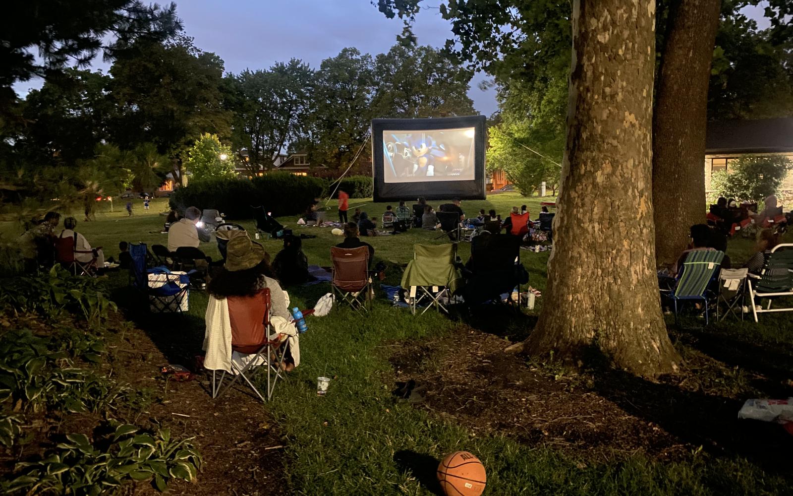 An image of people sitting in lawn chairs at the park watching a movie on a large screen