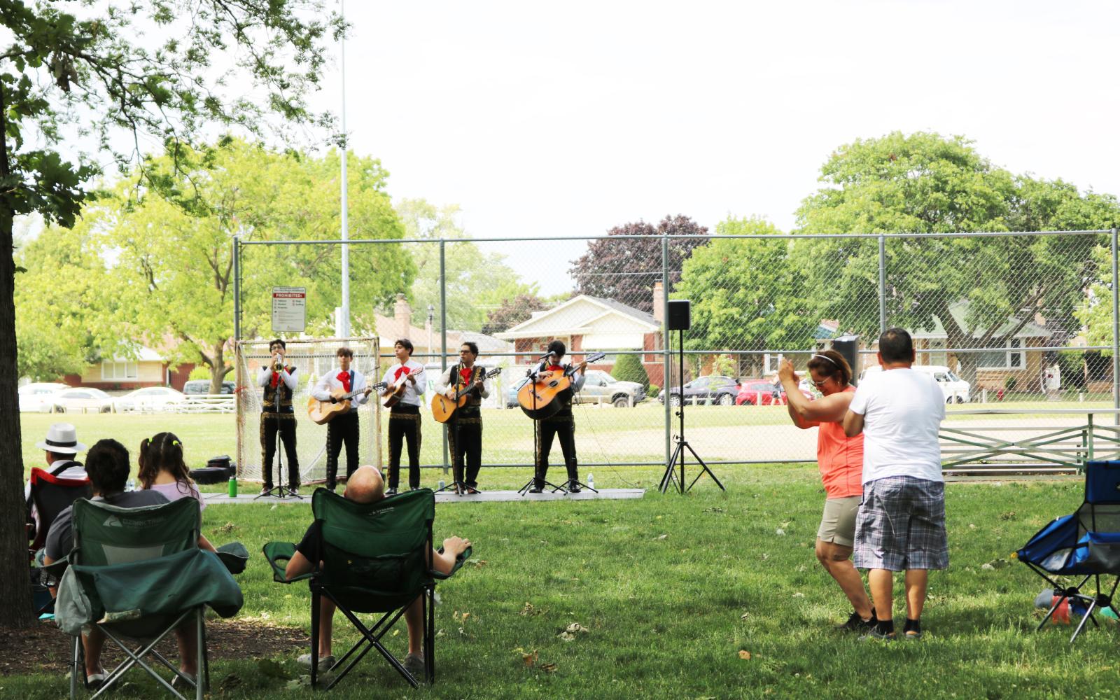 An image of a mariachi playing in the park with people dancing and watching in lawn chairs