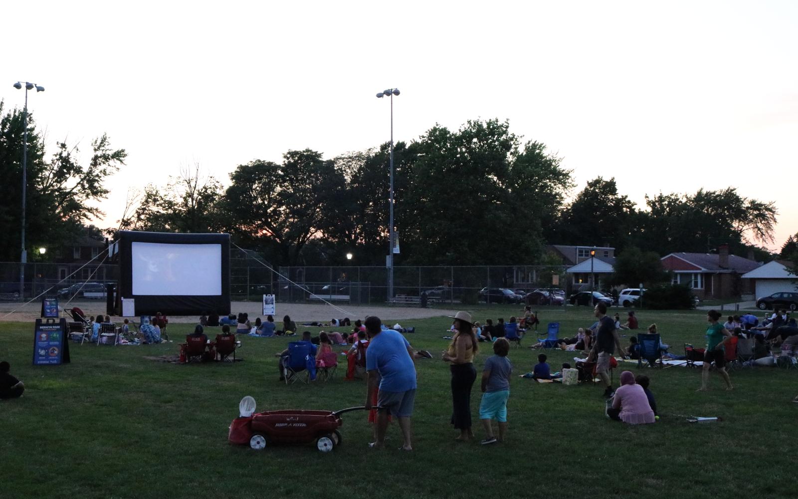 An image of community members sitting at the park and watching a movie near the baseball fields