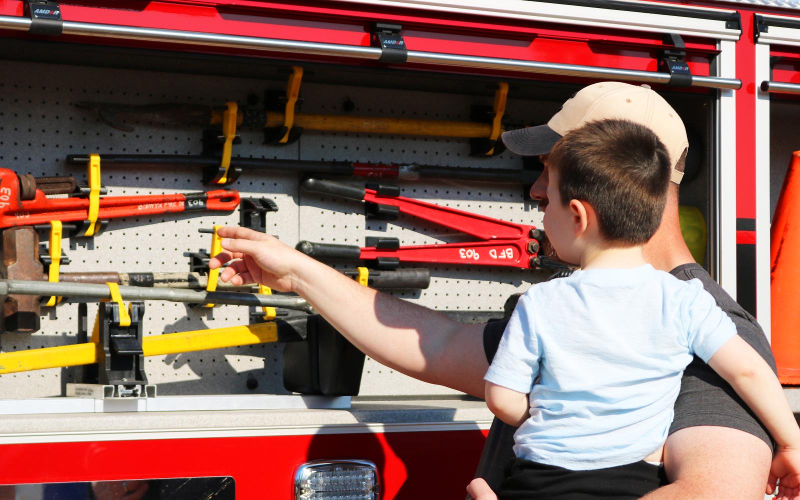 A parent holding a child and showing them the tools inside of a firetruck