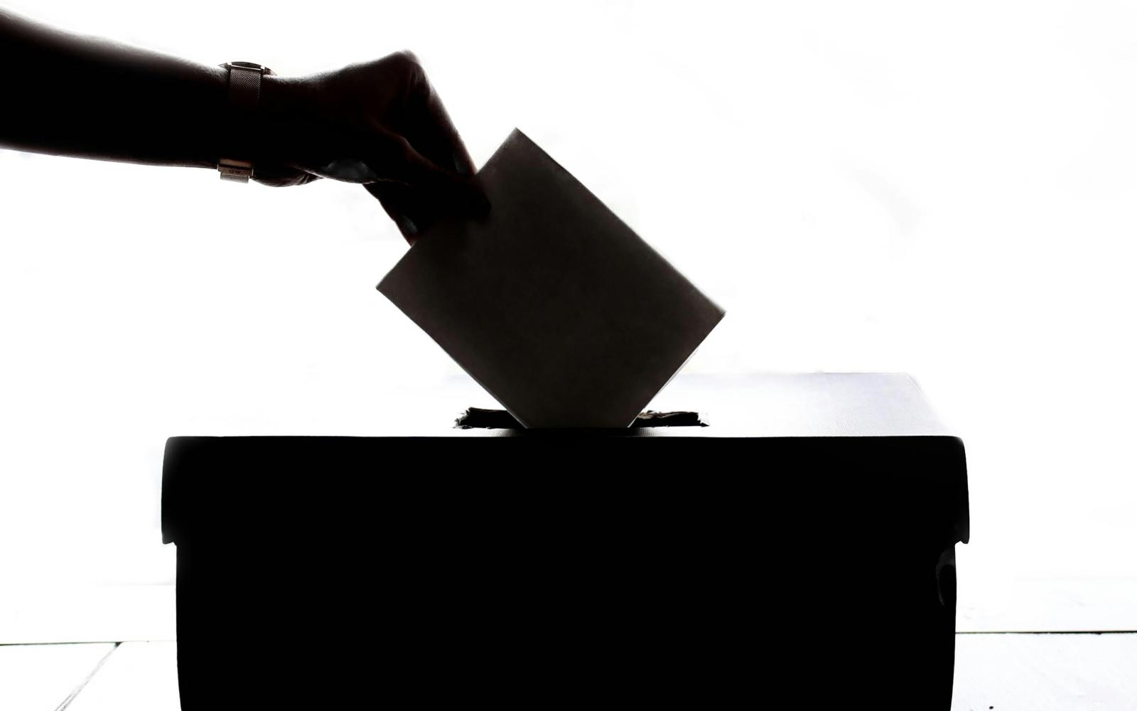A silhouette of a hand putting a folded paper into a ballot box