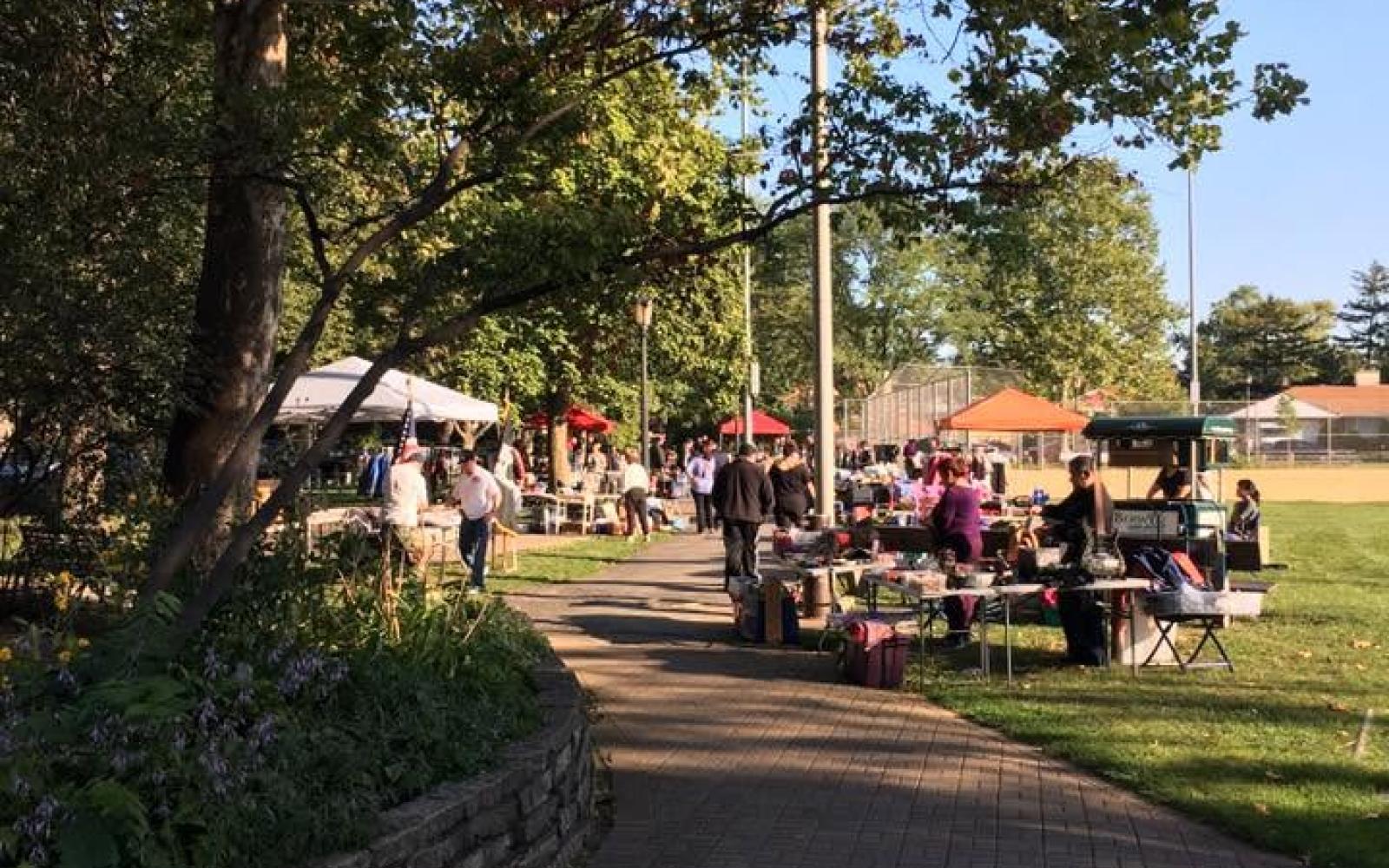 a pathway in Proksa Park leading towards the ball fields with people set up in tents on either side for the community garage sale