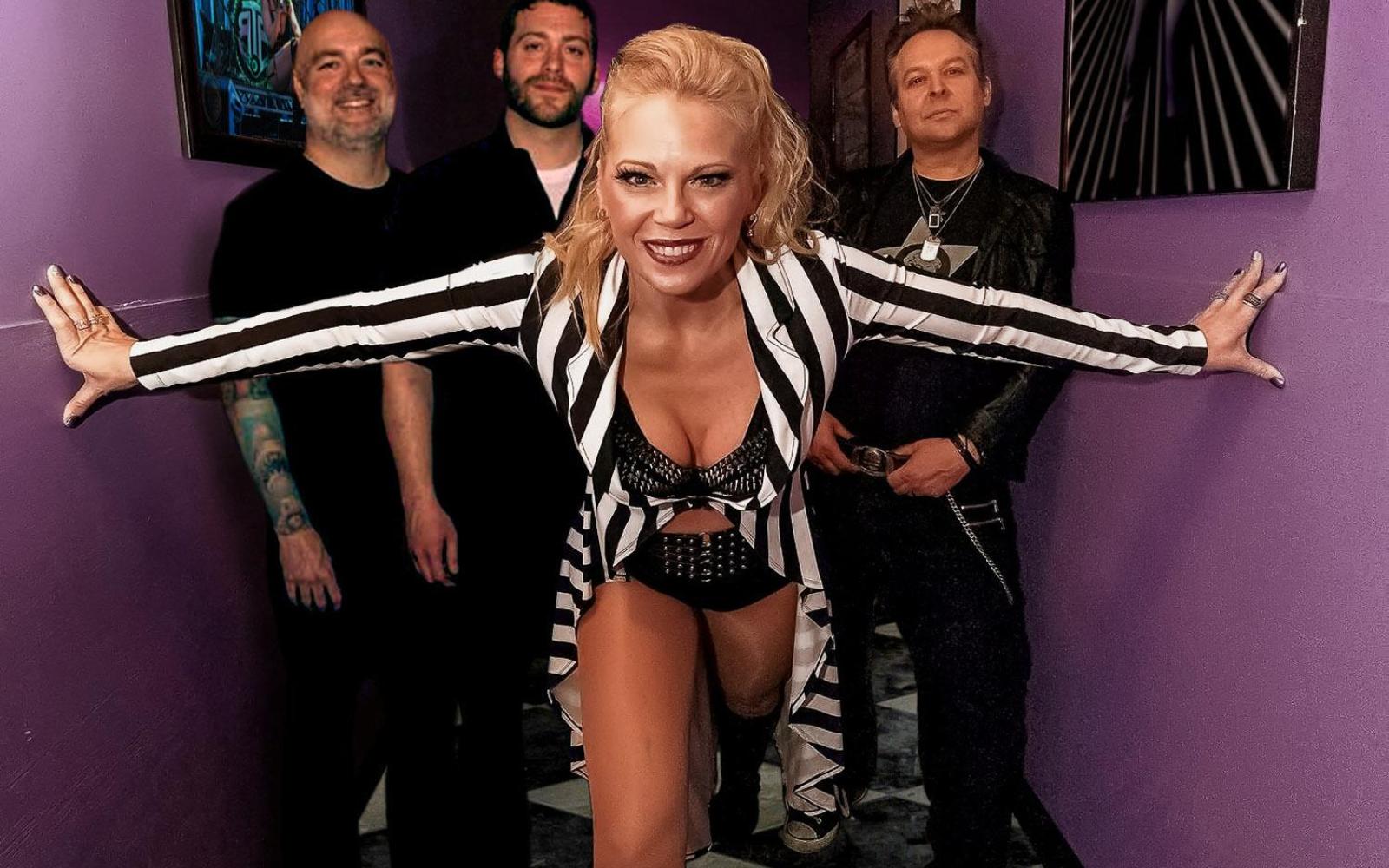 An image of a band in a hallway with purple walls. The female lead is in the front with her arms outstretched. Three male band members stand behind her.