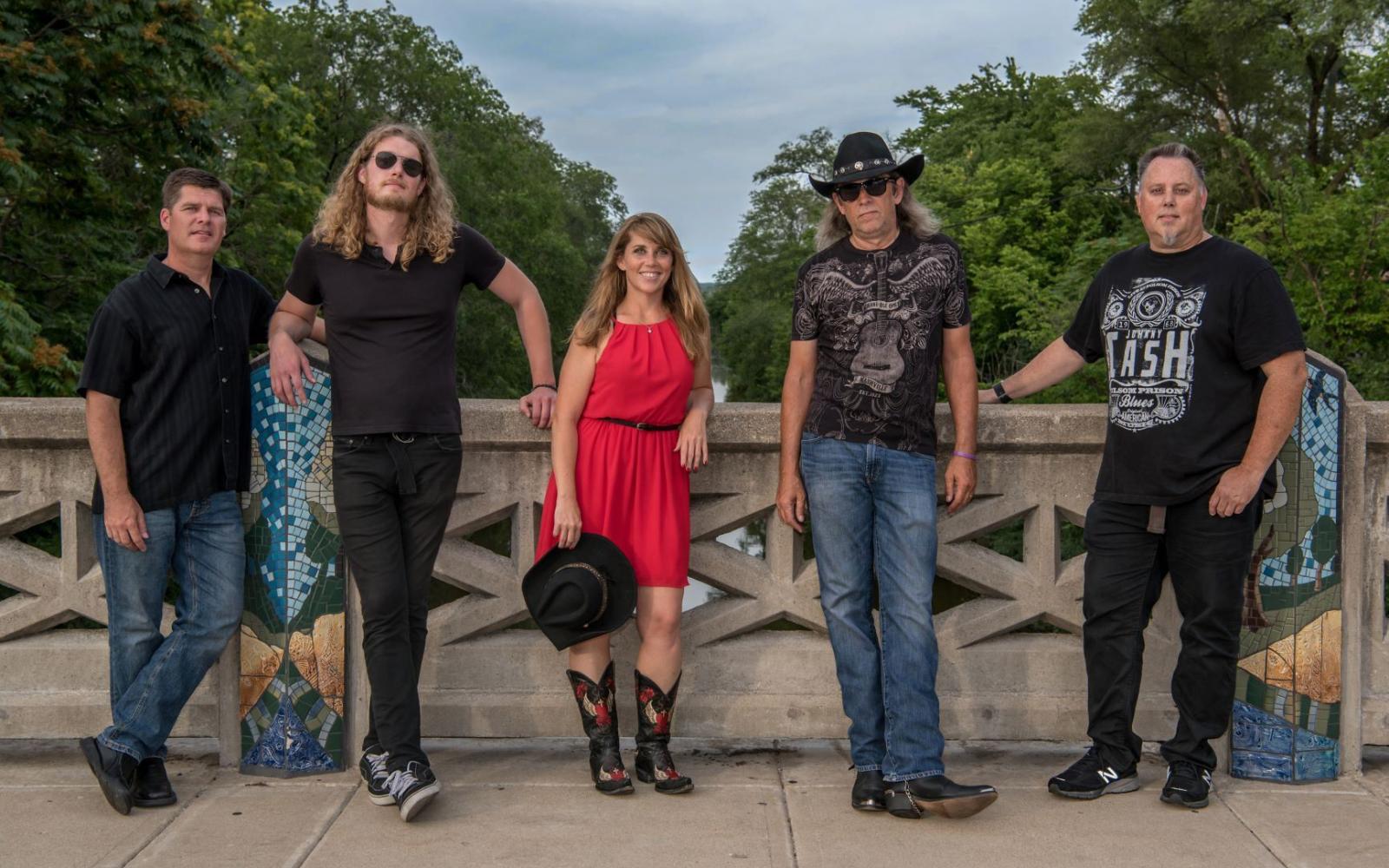 An image of the band "Wild Daisy" The picture depicts four men and one woman in the middle in a red dress standing on a bridge. 