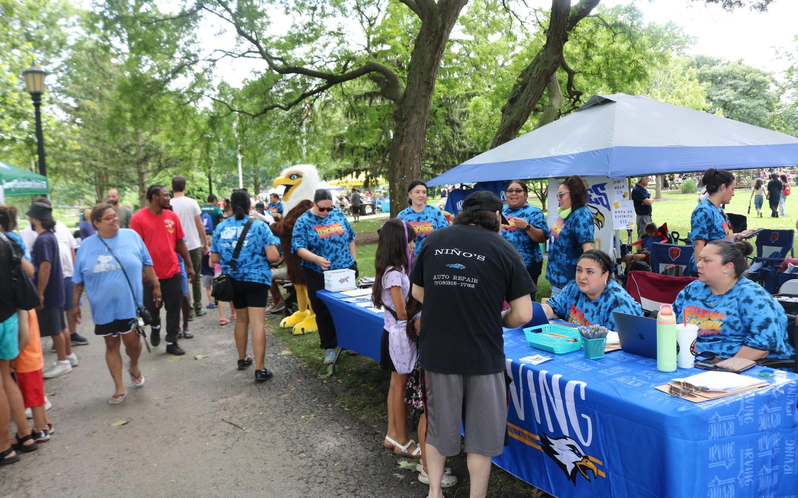 an image of a booth for a school PTO on a walkway with people gathering nearby. There is also an eagle mascot in the background.