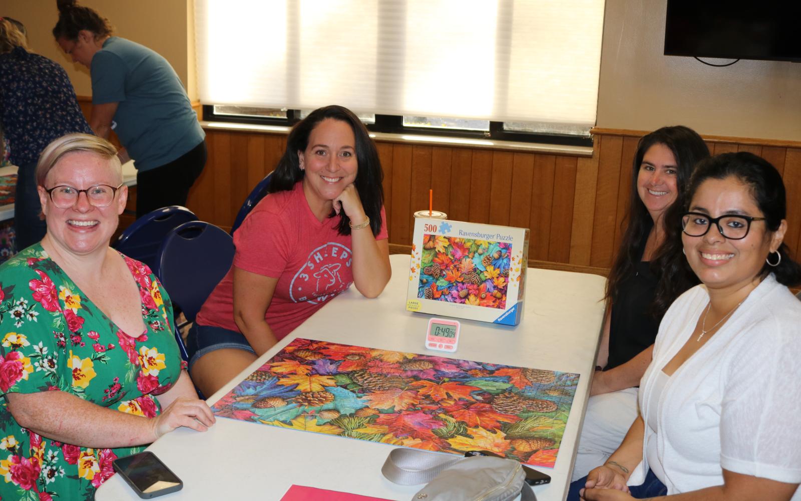 four women sitting around a white table with a completed puzzle on the table between them